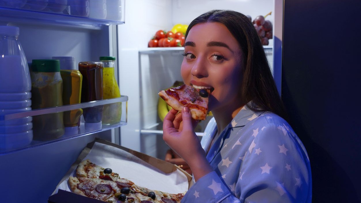 Young woman eating pizza near refrigerator in kitchen at night. Bad habit