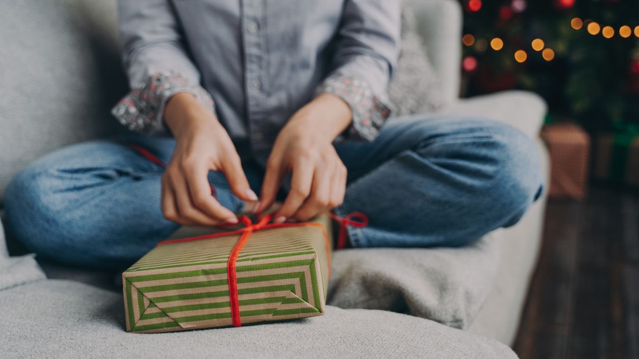 Cropped photo of woman opening christmas gift box at home, sitting on sofa near decorated xmas tree, female wrapping New Year present during winter holidays