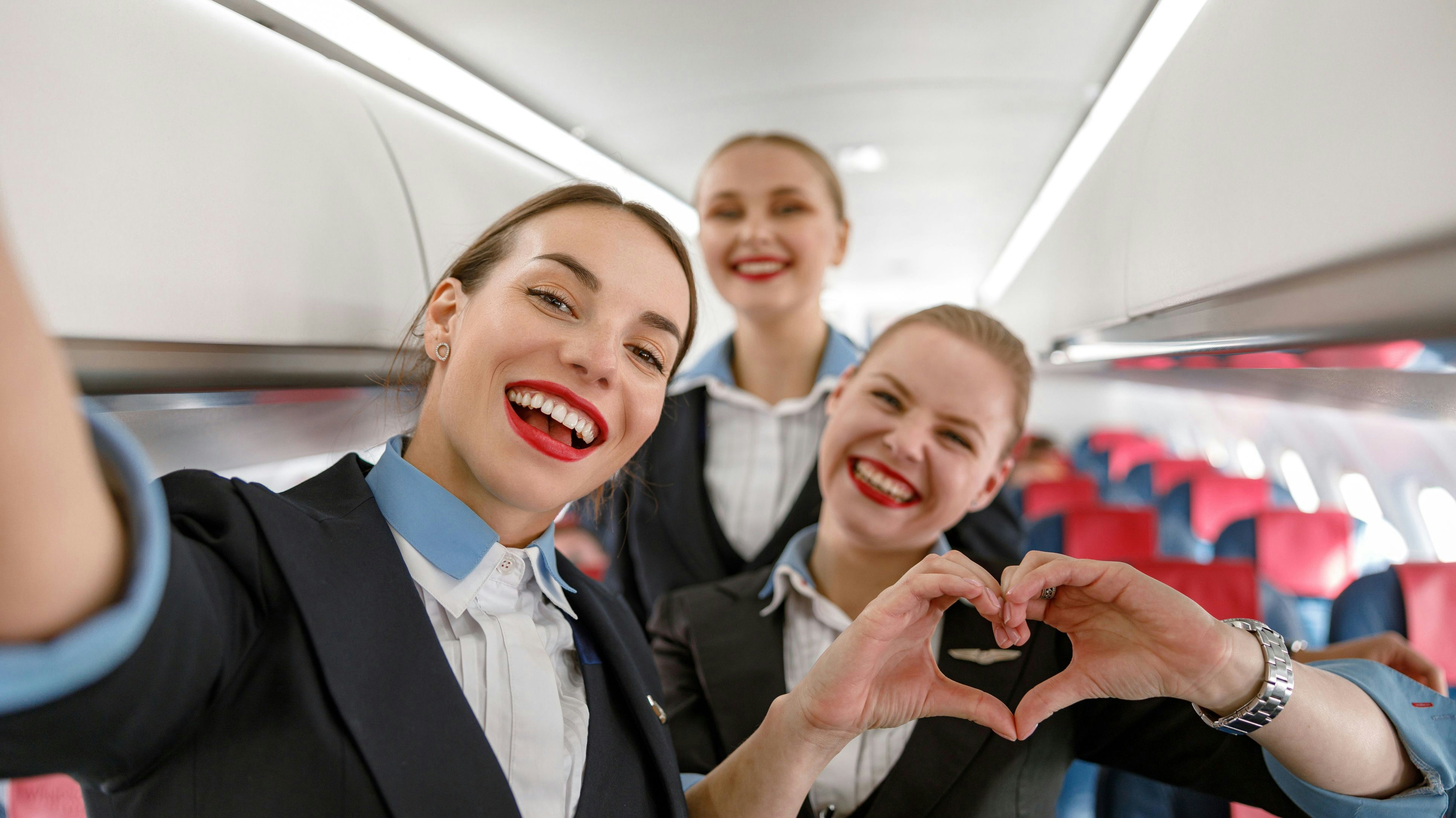 Joyful woman stewardess doing heart symbol with hands and smiling while standing with colleagues in aircraft cabin
