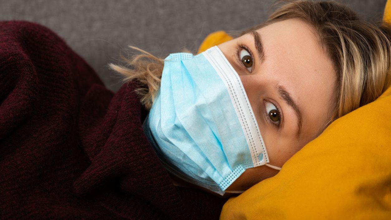 Young woman with coronavirus is having headache and fever, lying on the sofa wrapped in a balnket