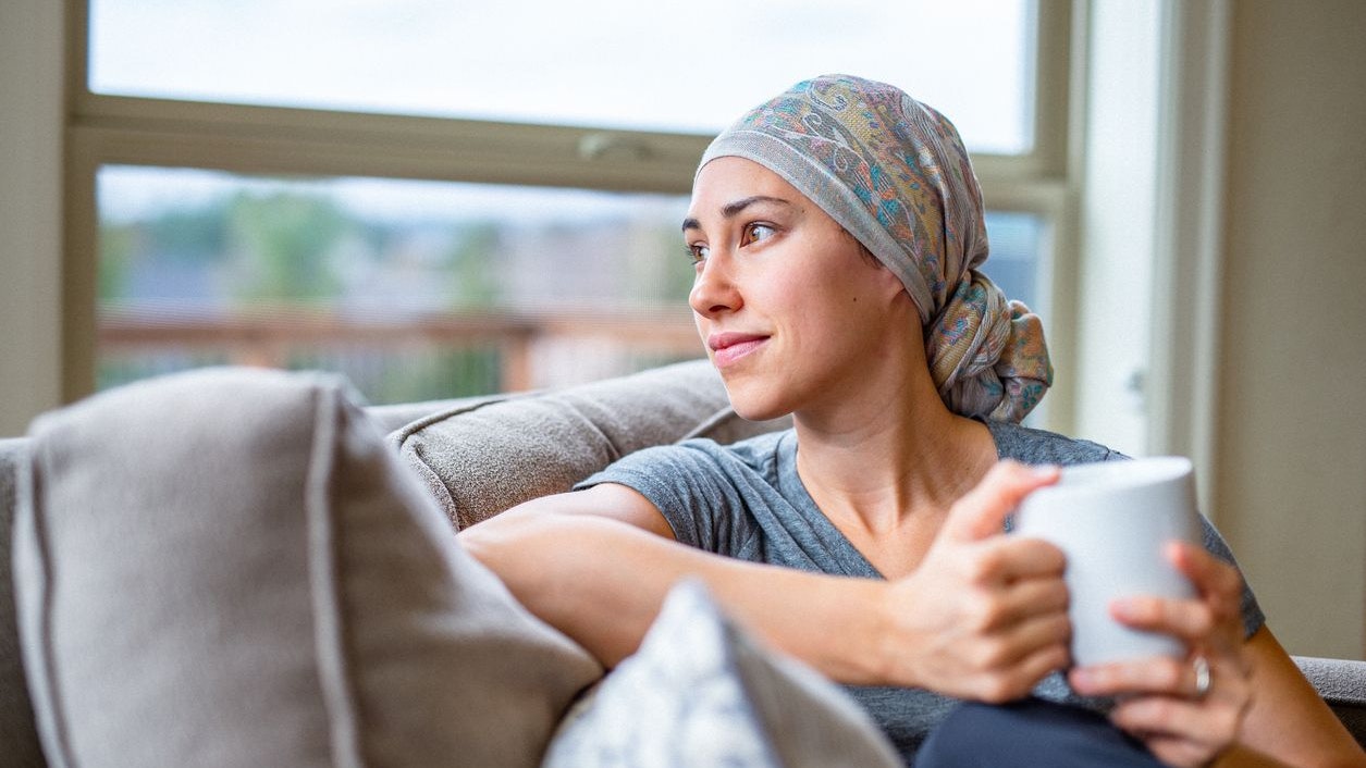 A brave and courageous woman of Eurasian descent wearing a headscarf and living with cancer relaxes on the couch at home, smiling while holding a coffee mug.