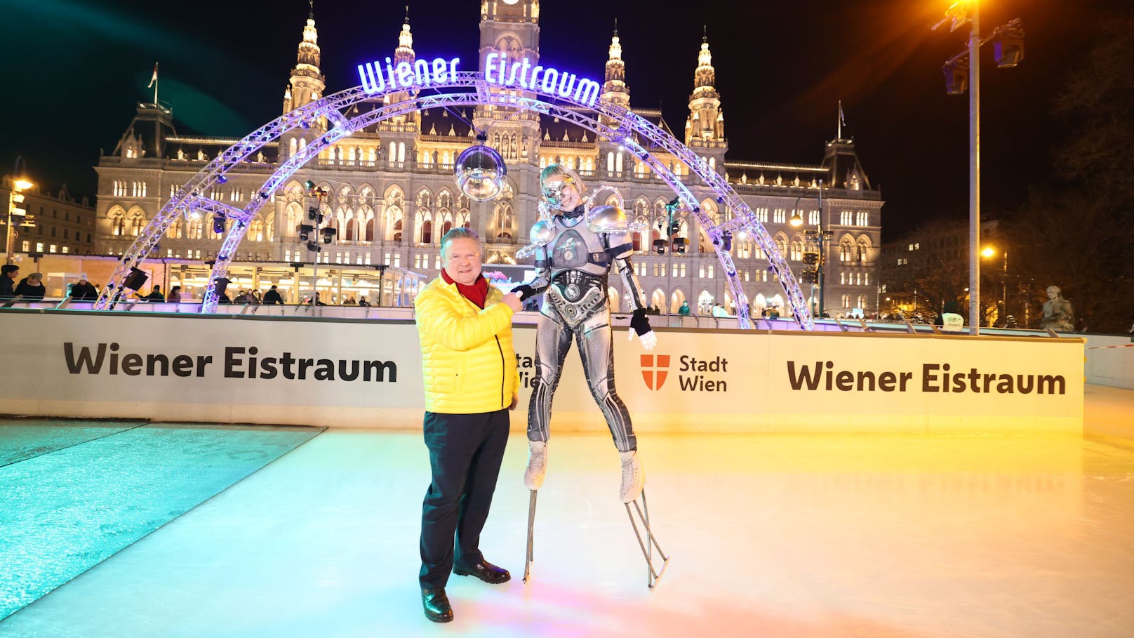 Ein Wintertraum wird wahr – Bürgermeister Ludwig und ein futuristischer Eisläufer vor der märchenhaften Kulisse des Rathauses.