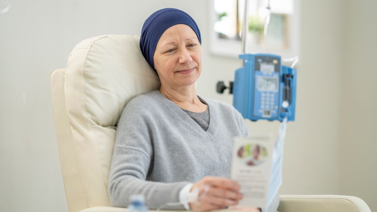 A woman sits in a chair as she receives her Chemotherapy treatment intravenously.  She is dressed comfortably and has a headscarf on to keep her warm as she reads a pamphlet on what to expect during treatment.
