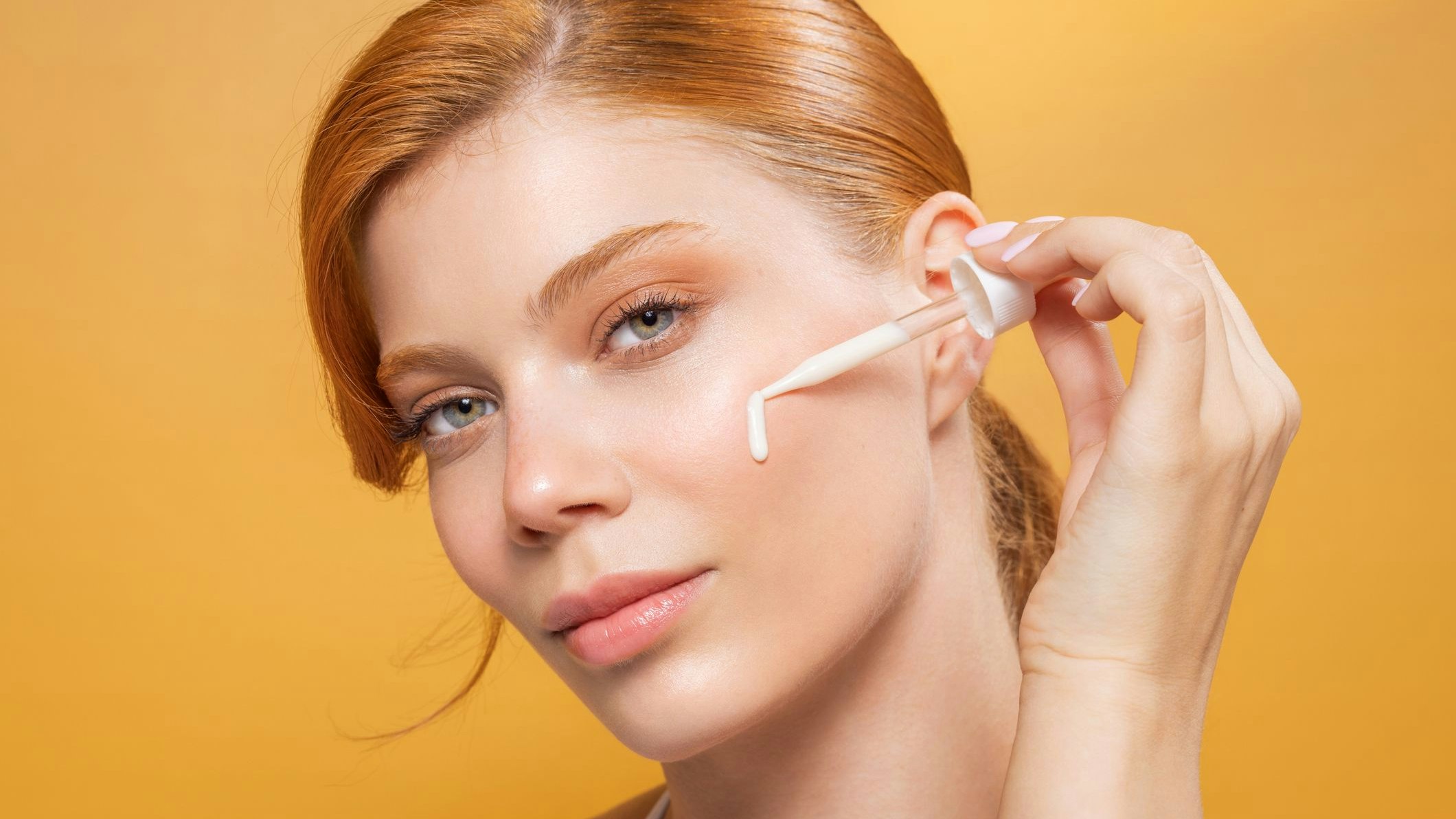 Studio shot of a beautiful redhead woman with perfect skin, applying facial serum on skin.
