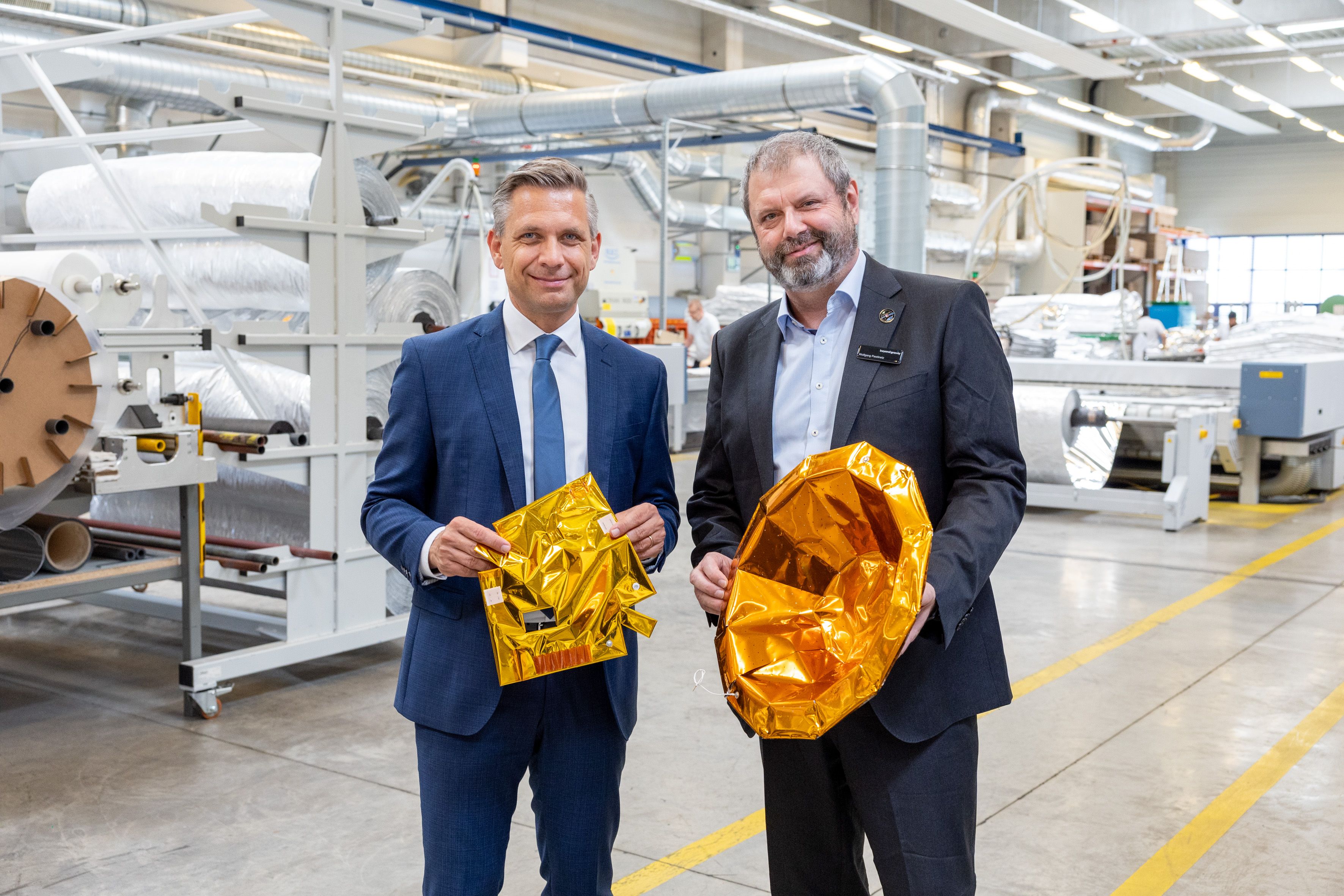Wirtschaftsminister Wolfgang Hattmannsdorfer und Wolfgang Pawlinetz, Geschäftsführer Beyond Gravity Austria, mit goldfarbener Thermalisolation für Satelliten.