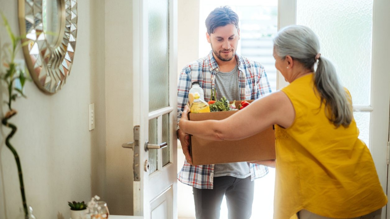 Woman delivering a box of groceries to a man at his home entrance. Concept of food delivery, assistance, and community support.