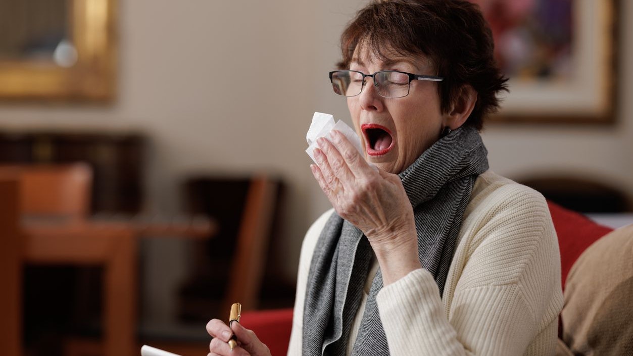 Senior woman indoors holding a tissue, about to sneeze with eyes closed. She is also holding a pen and note pad.