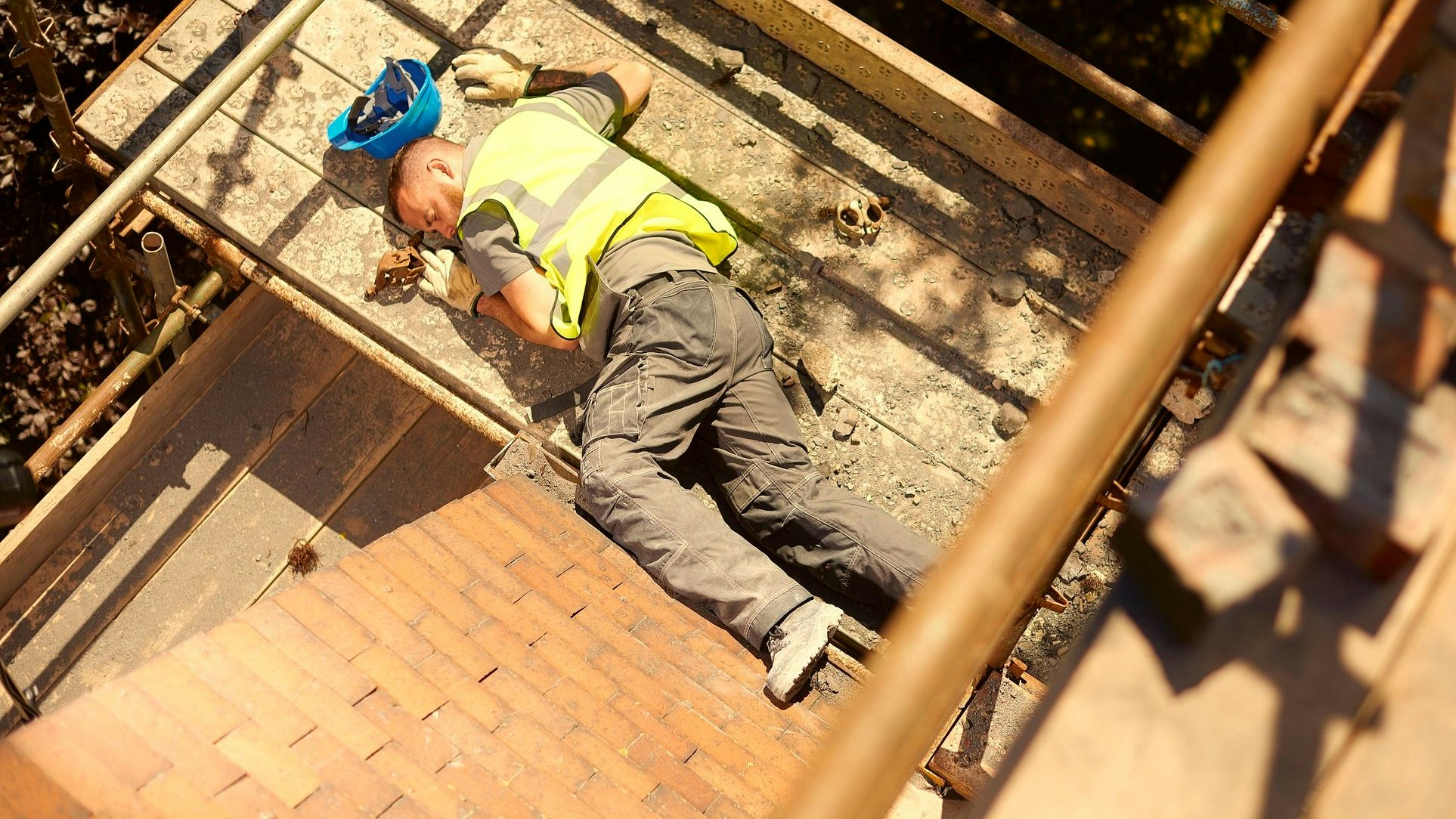 a builder or roofer has fallen from the top elevation of scaffold on a construction site and landed on the next level . He is grimacing in pain .