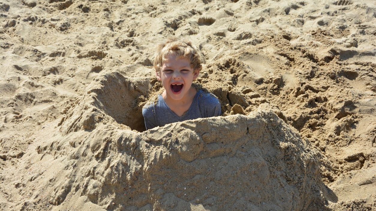 Boy has dug himself in in sand and smiles forwards
