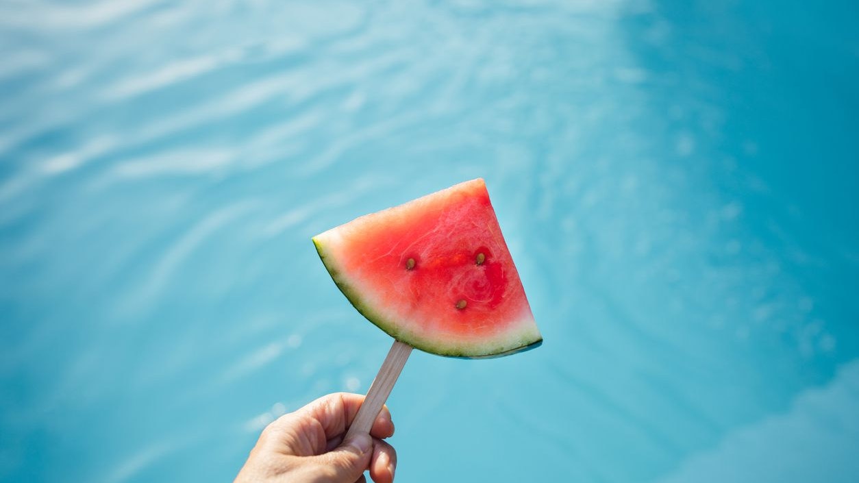 A close-up shot of a hand holding a watermelon slice on a stick with a clear pool in the background