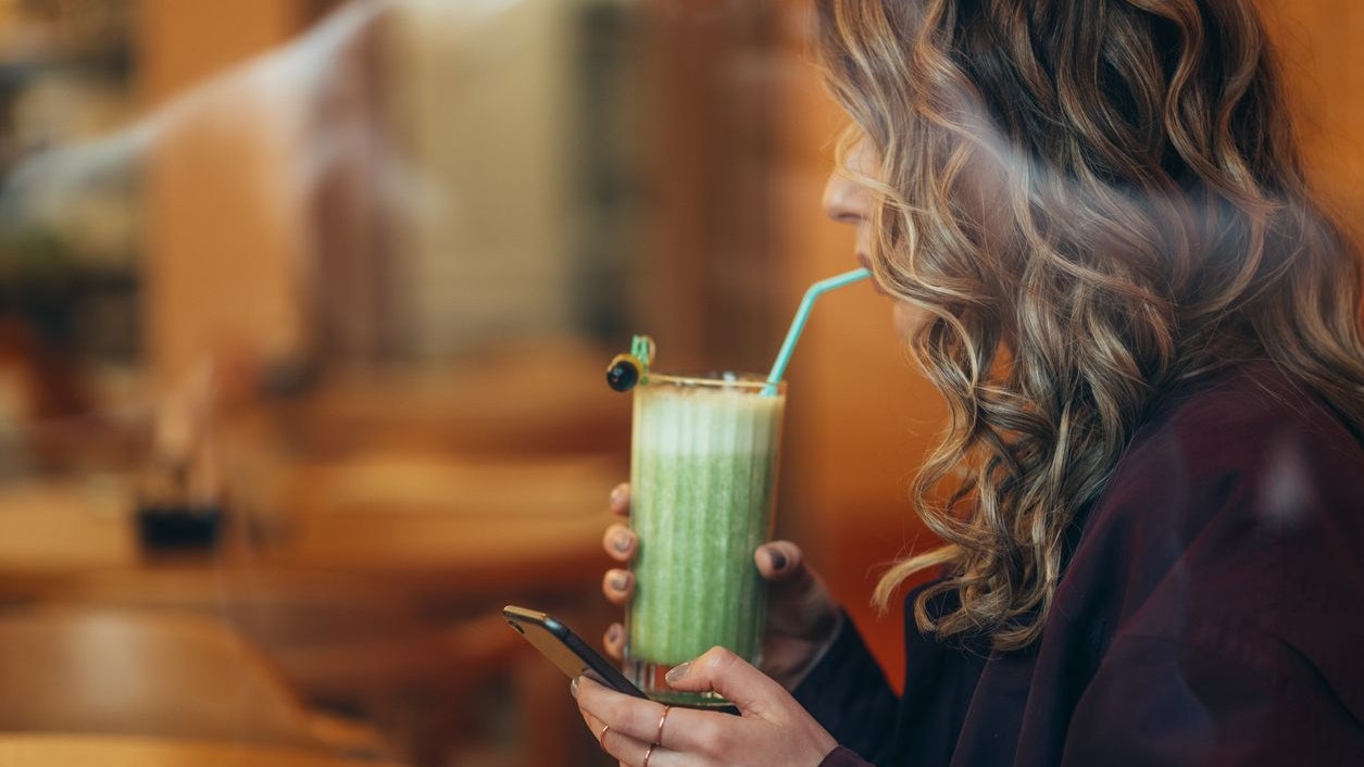 Beautiful young woman drinking matcha latte coffee and using smartphone in a cafe