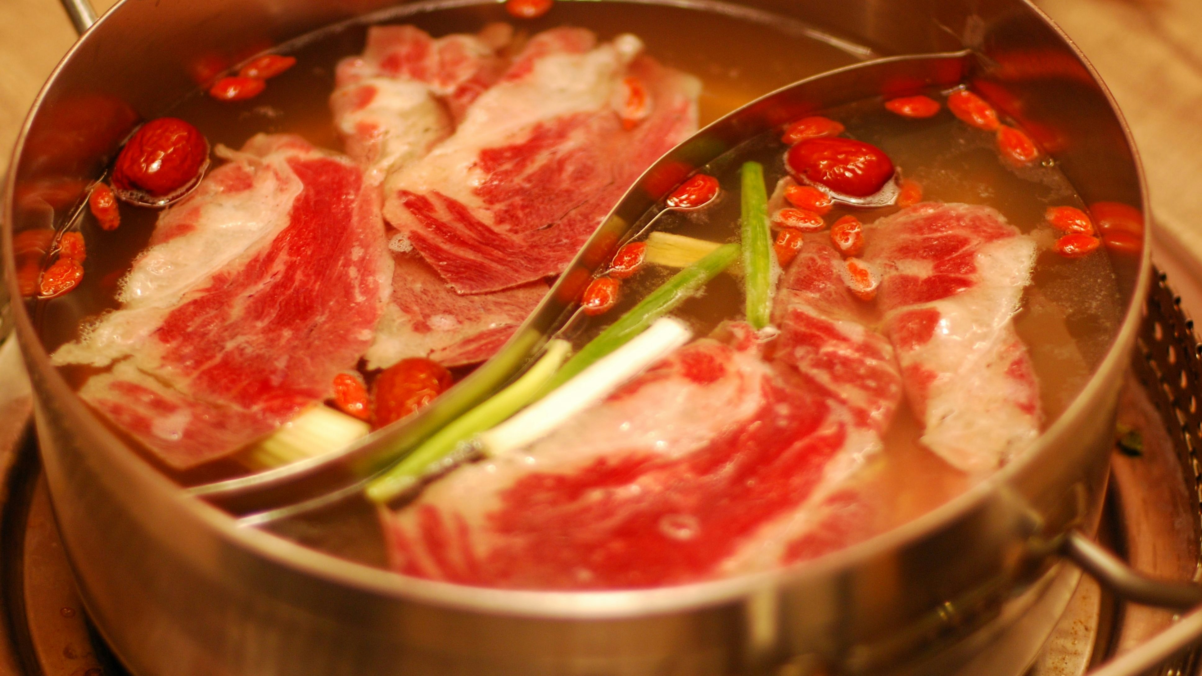Close-up of a hot pot with raw beef slices, goji berries, and green onions simmering in broth.