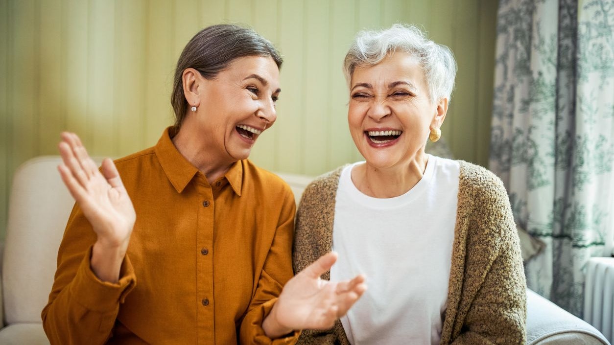 Elderly sisters watching comedy together indoors, laughing. Portrait of two beautiful Caucasian women relaxing at home, sitting on sofa, opening mouth wide opened, enjoying weekend, having fun