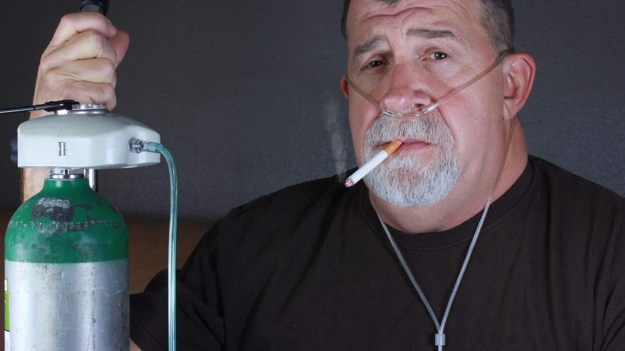 A adult man dangerously smokes a cigarette while wearing an oxygen cannula while holding on to an oxygen gas tank.