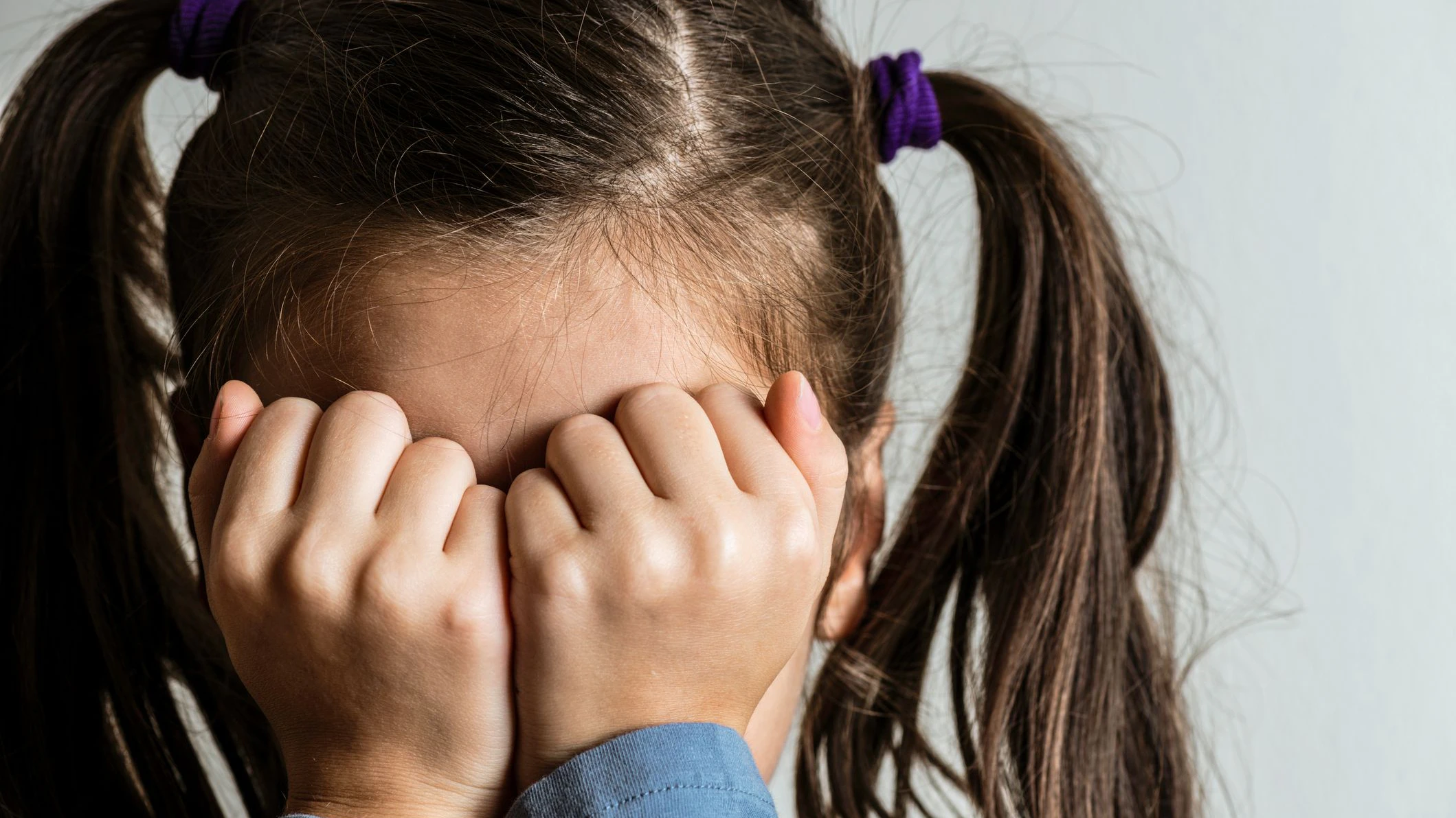 Little crying caucasian girl with double ponytail who is hiding her face with her hands. Representing child abuse and domestic violence.
