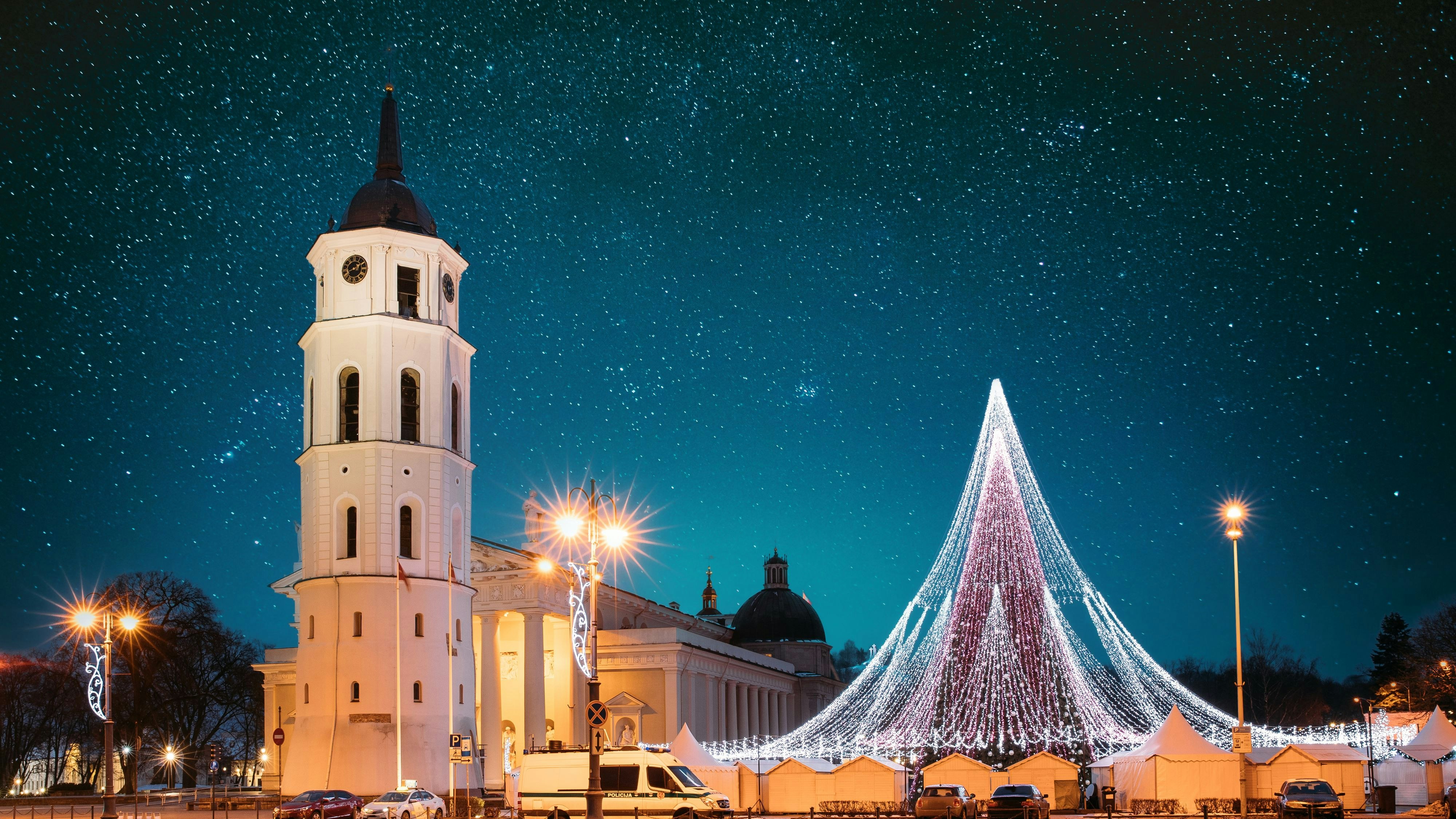 Vilnius, Lithuania. Amazing Bold Bright Blue Starry Sky Gradient Above Christmas Tree On Background Bell Tower Belfry Of Vilnius Cathedral At Square In Evening New Year Christmas Xmas Illuminations.