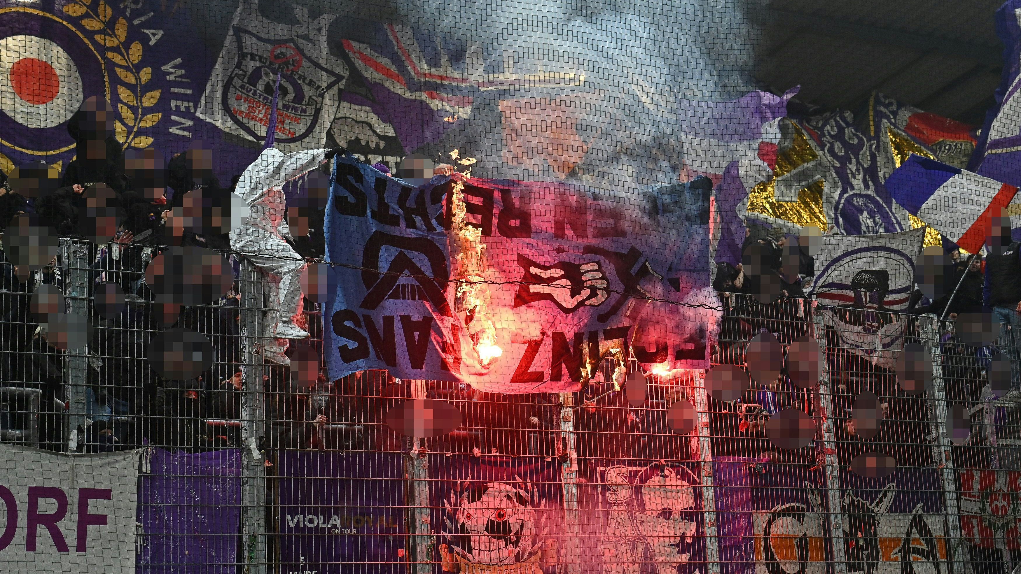 Das Foto zeigt zwei Anhänger von Austria Wien beim Match in Linz. Sie verbrennen dabei ein altes Transparent des FC Linz von 1994. Darauf steht: "FC Linz Fans gegen Rechts", daneben ist eine Faust, die ein Hakenkreuz zerschlägt.
