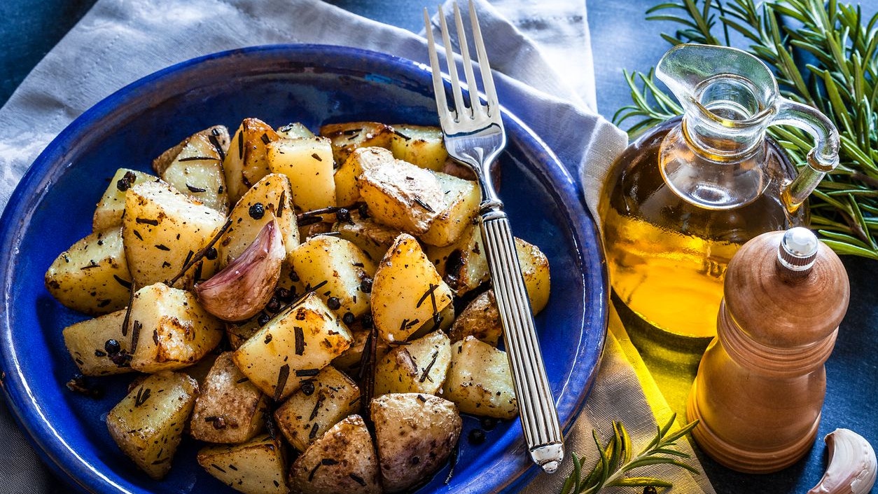 High angle view of a blue plate filled with roasted potatoes shot on rustic wooden table. The plate is at the bottom of an horizontal frame placed on a bluish textile napkin and an olive oil bottle and a pepper shaker are at the top placed directly on the table. Predominant colors are brown and yellow. Low key DSRL studio photo taken with Canon EOS 5D Mk II and Canon EF 100mm f/2.8L Macro IS USM