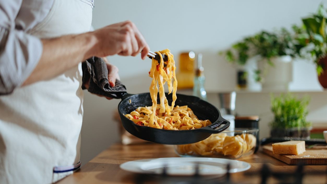 An anonymous chef in apron picking up tagliatelle with serving tongs from the frying pan and serving it on the plate.