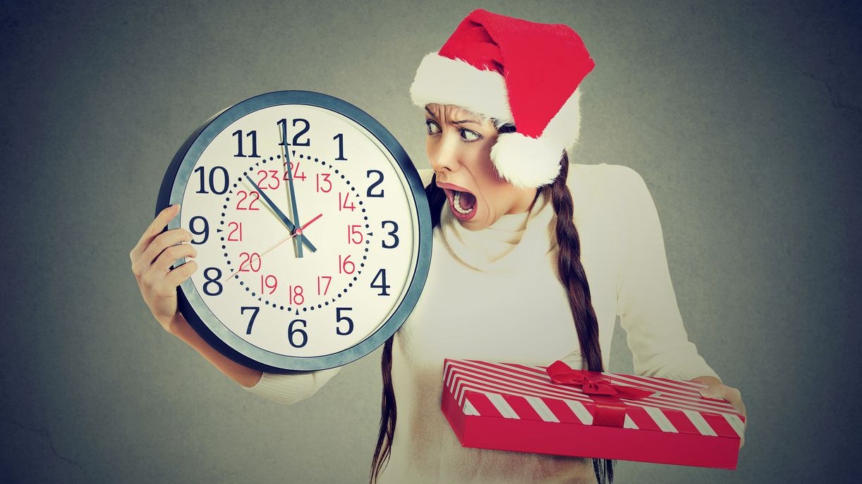Closeup portrait worried stressed in a hurry young woman wearing red santa claus hat holding clock gift box isolated gray background. Emotion, funny face expression, last minute christmas shopping