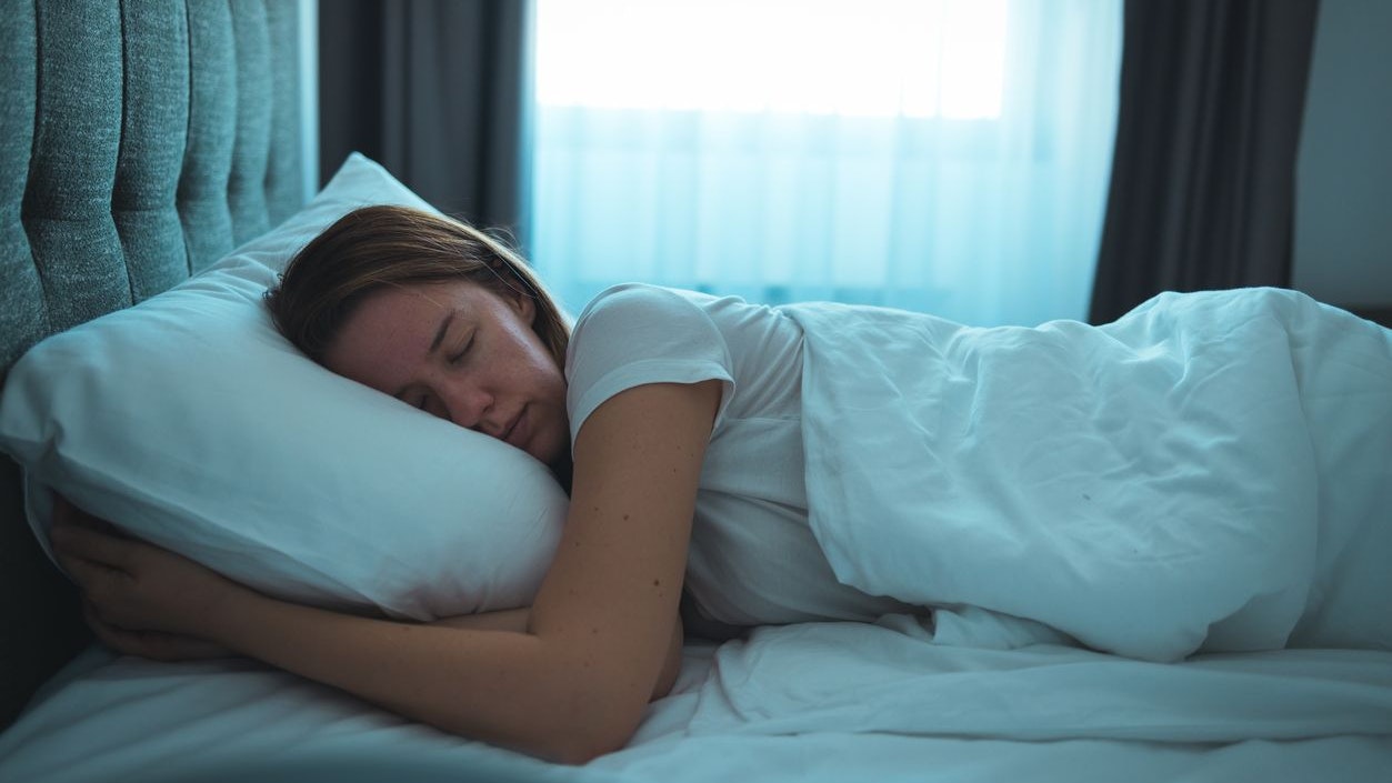 Side view of young beautiful woman dreaming in bed and relaxing at night. High angle view of woman with closed eyes sleeping well at home in the dark. Beautiful girl sleeping peacefully under white blanket in her bedroom at late in night.