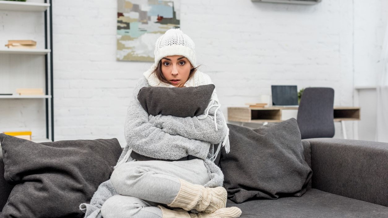 freezed young woman in warm clothes sitting on couch and hugging cushion at home