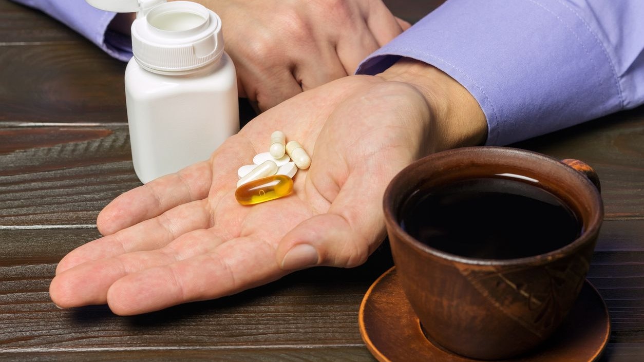 Male hand holds vitamin complex and dietary supplement, capsules of omega 3, glucosamine, calcium pills, cup of black coffee on dark wooden table.