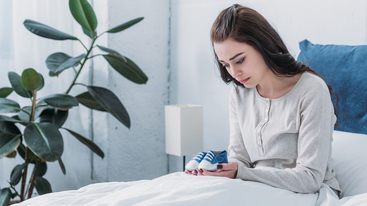 depressed woman holding baby shoes while lying in bed
