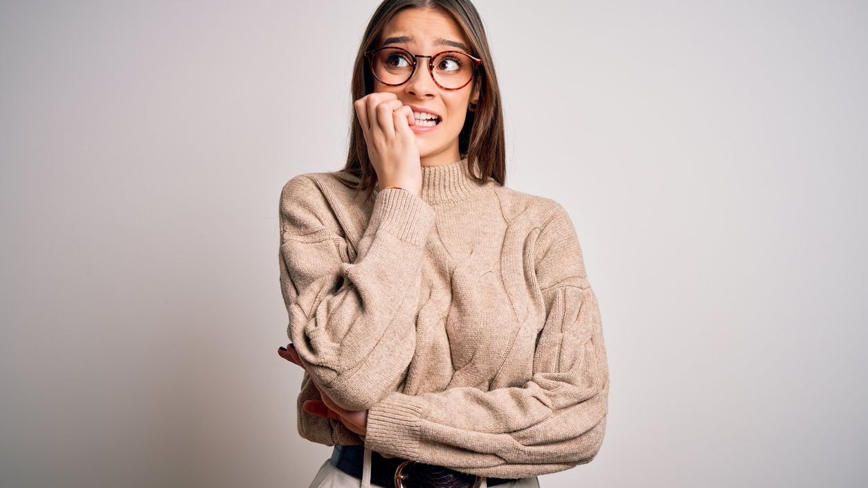 Young beautiful brunette businesswoman wearing casual sweater and glasses standing looking stressed and nervous with hands on mouth biting nails. Anxiety problem.