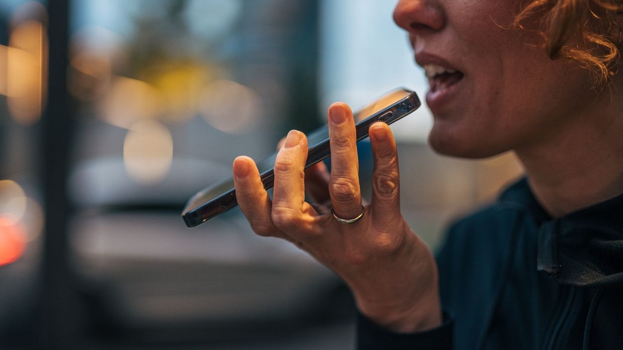 Woman sending a voice message on smartphone in a nighttime city scene, casually holding device near mouth as she speaks, modern mobile communication and social media use