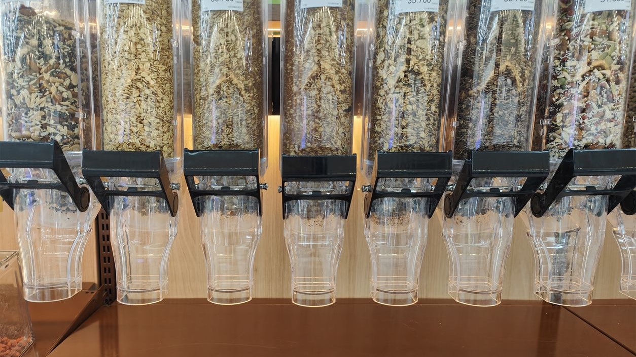 close-up shot of transparent bulk dispensers filled with different varieties of granola, oats, and cereal blends in a grocery store. Labels and prices are visible, offering natural and healthy food choices for customers.  Concept: bulk food, granola, healthy eating, oats, cereal dispensers, zero waste shopping, supermarket, natural food, bulk store, sustainable lifestyle, plastic-free shopping, eco friendly