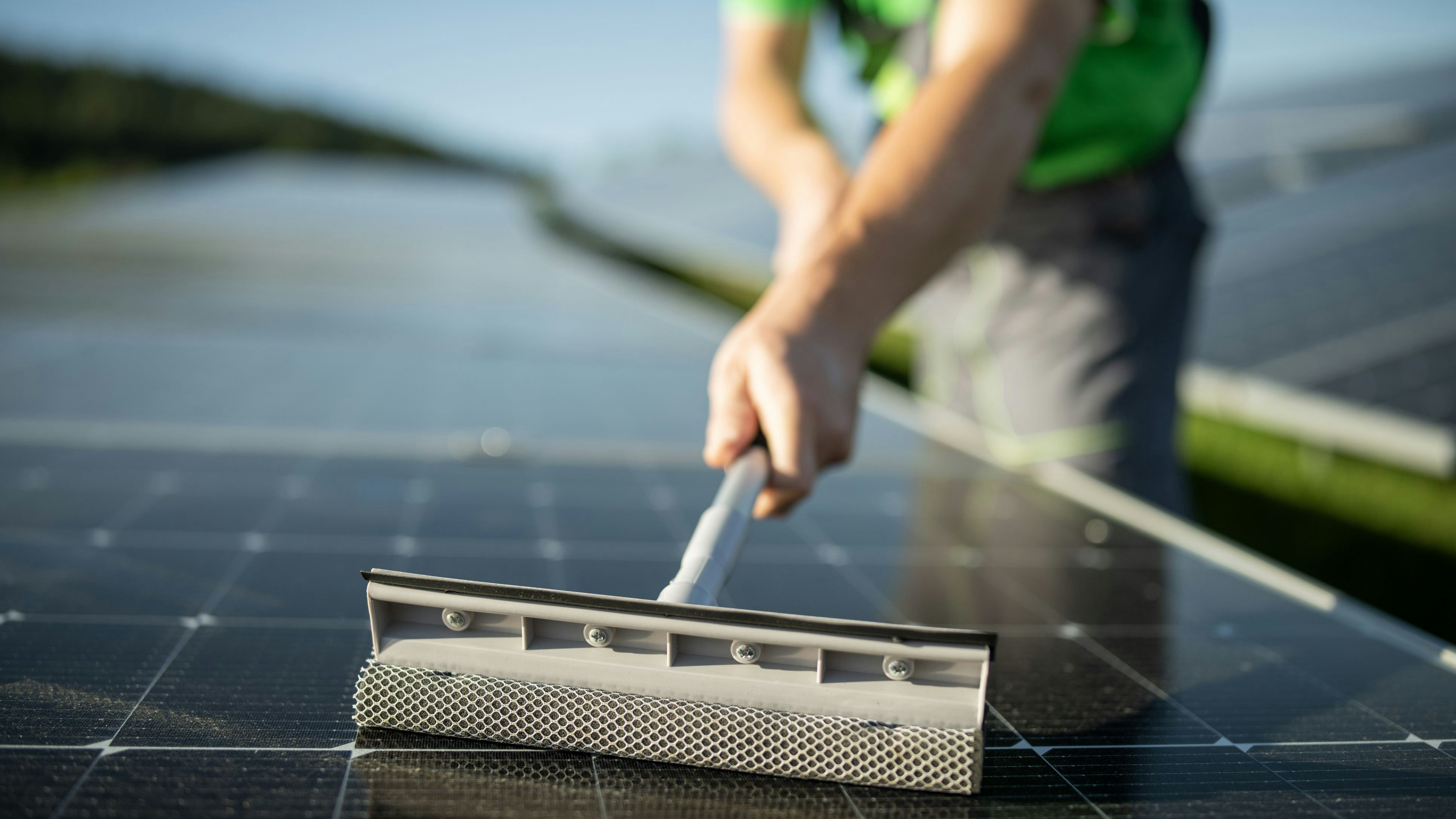 Close up of unrecognisable worker in green clothes cleaning solar panels with tool.