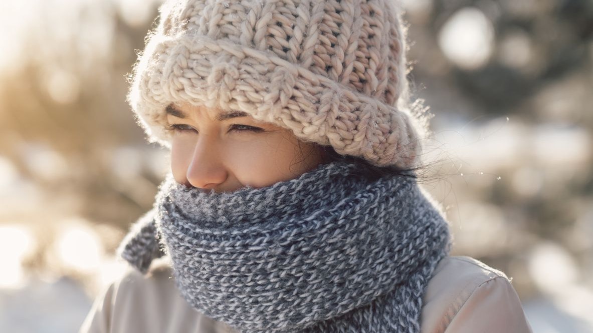 Portrait of funny woman in casual winter clothes hat scarf and coat. Young woman suffering in a cold winter outdoors. Cute funny mixed race Caucasian Asian woman close-up standing against winter background looking away.