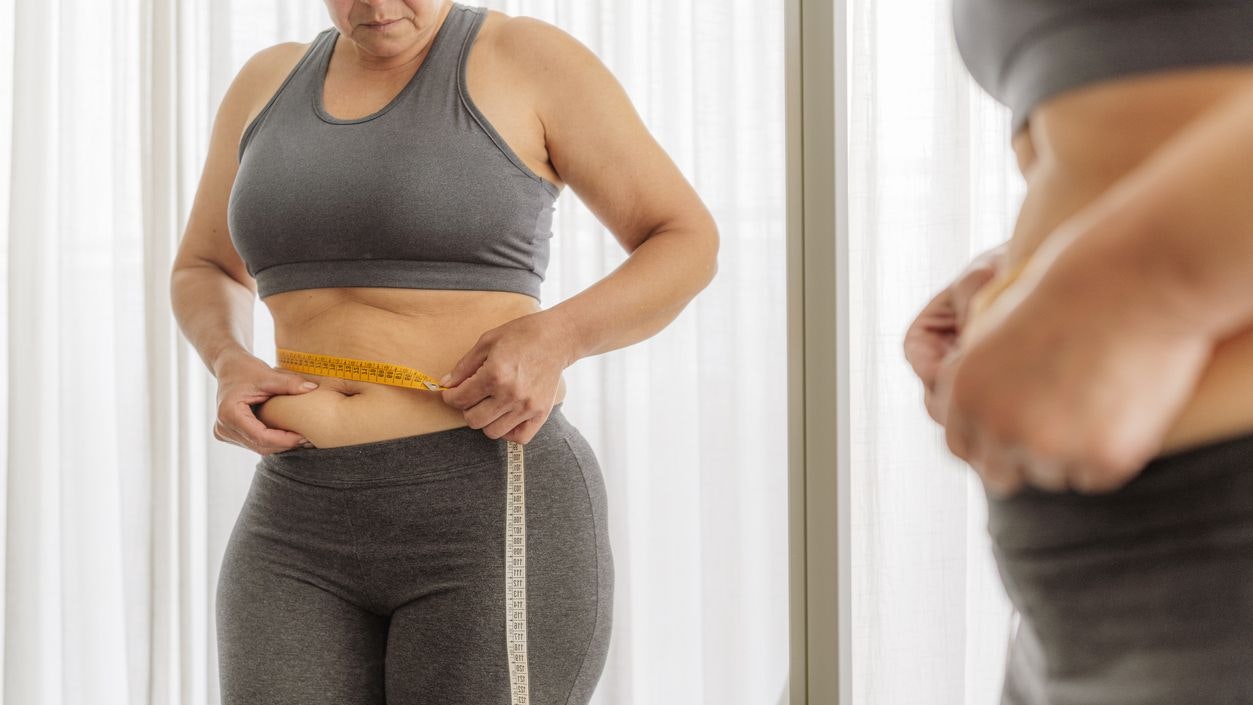 Mature overweight woman in sports clothing measuring her waist and squeezing her belly fat in front of the mirror