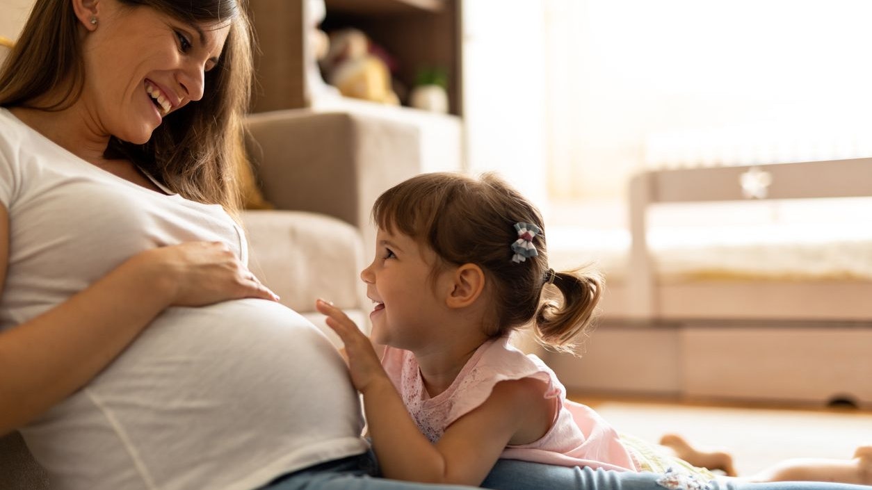 Happy small girl holding hands on mothers belly and listening baby's heartbeat. Innocent girl Touching Abdomen Of Pregnant Mother At Home