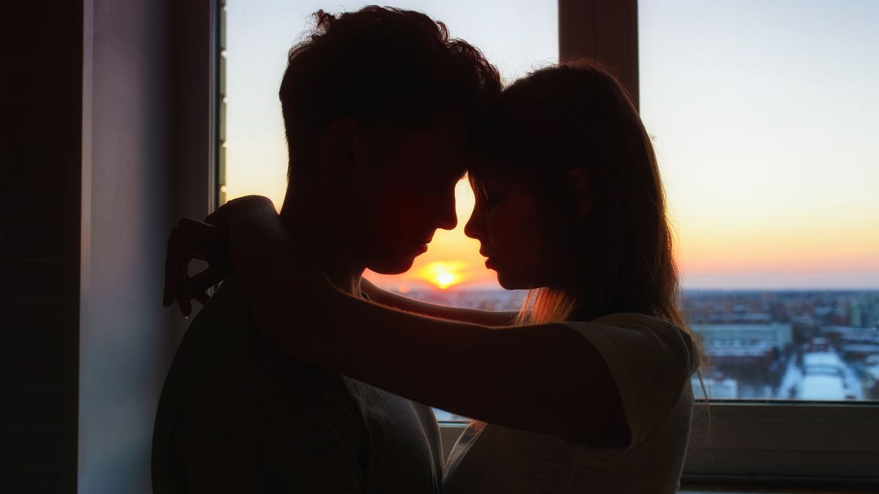 Couple in love, profile silhouettes close to each other, beautiful sunset in window at background