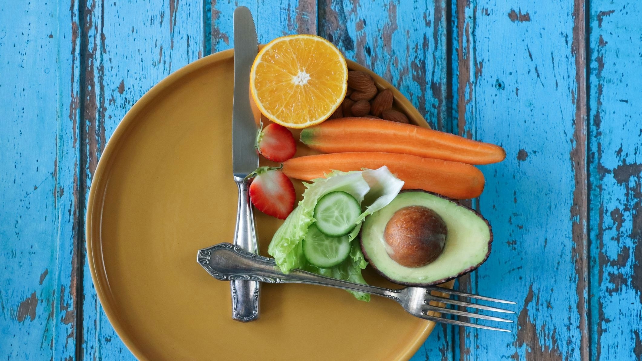 Stock photo showing an intermittent fasting concept depicted by a plate and cutlery forming a clock face and hands with the smallest section on the plate being filled with a pile of almonds, half an orange, strawberry cut in two, a sliced carrot, lettuce leaves, slices of cucumber and half an avocado displaying it's large seed.