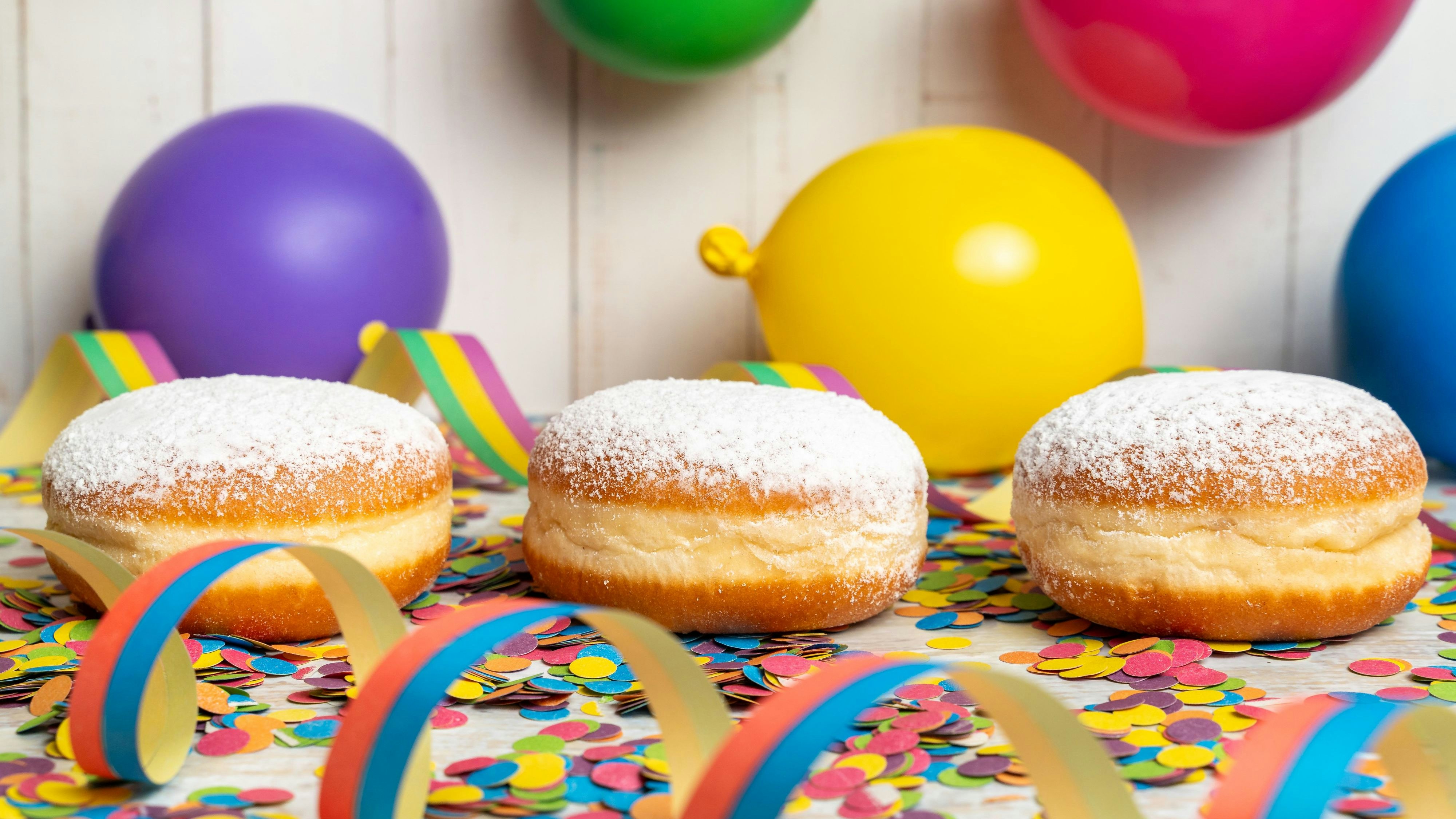 Drei klassische Bäcker-Krapfen mit Puderzucker liegen zwischen Konfetti und Luftballons. Das süße Gebäck steht für Fasching, Karneval und traditionelle Feiern aus der Bäckerei.