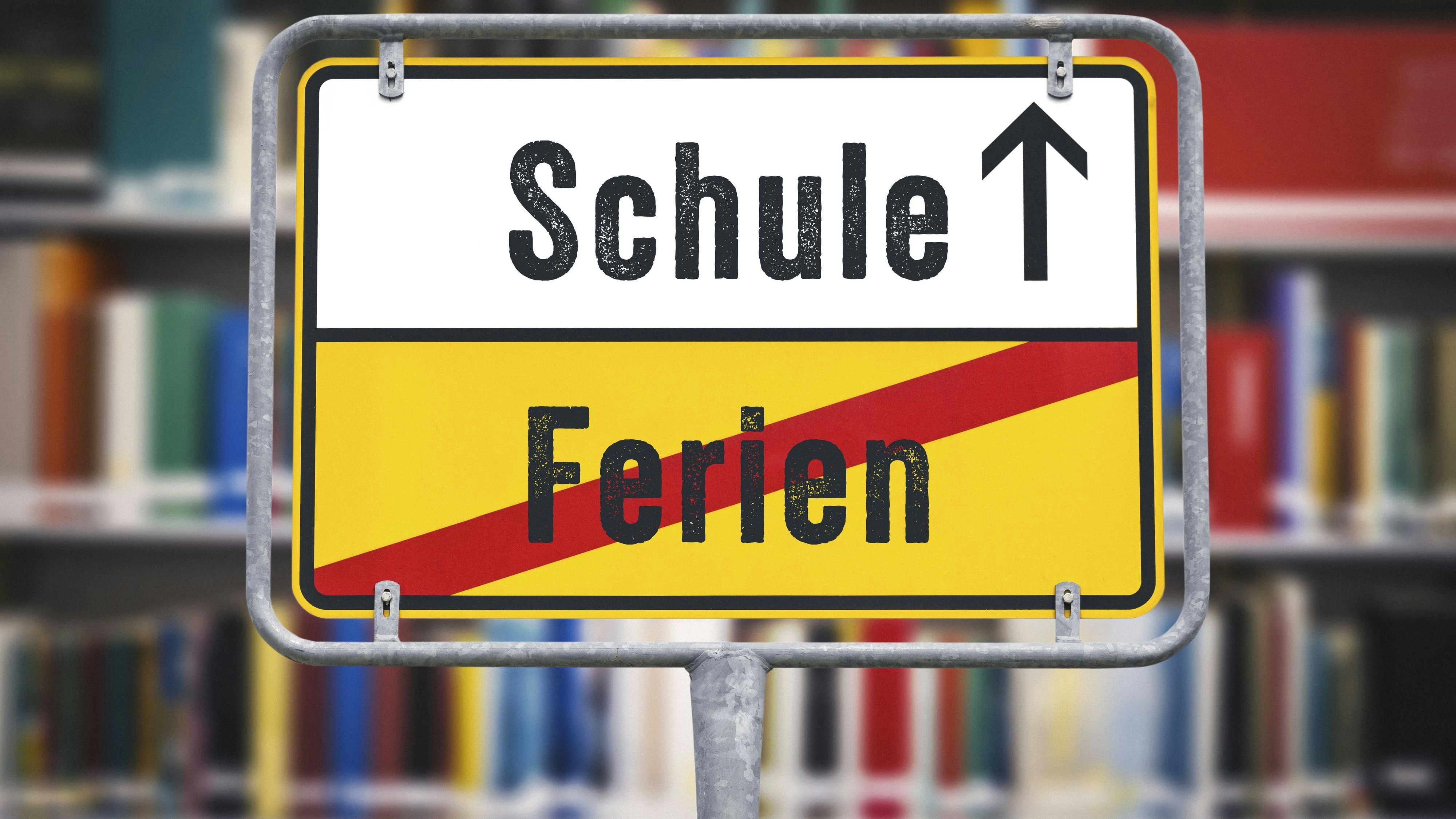 Ortsschild in einer Bücherrei oder einem Klassenzimmer mit dem Wort Ferien durchgesrichen und einem Pfeil in Richtung Schule. Symbolbild Schulanfang für Kinder in der Schule FOTOMONTAGE