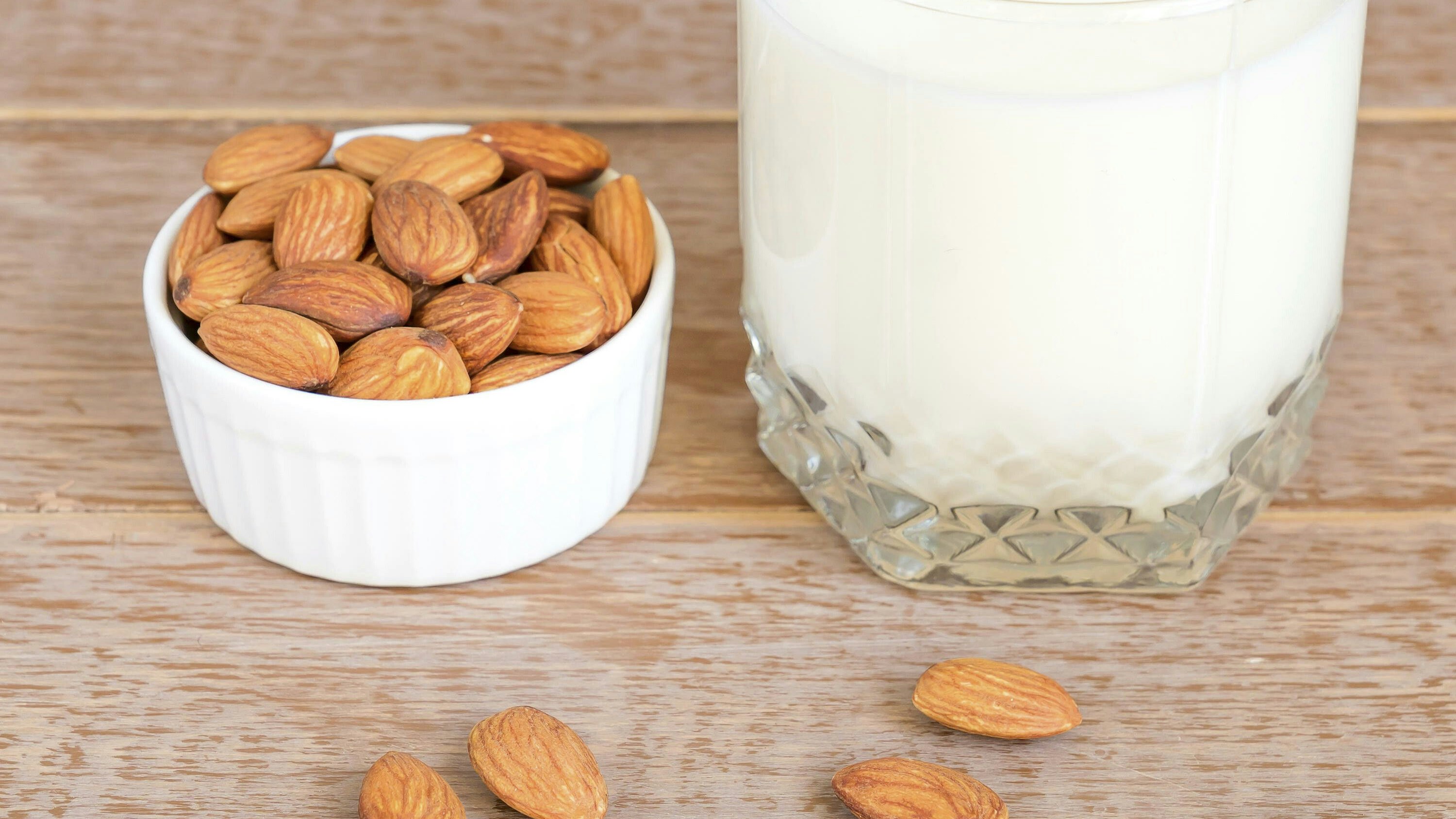 Homemade Almond milk in a glass and nuts in white porcelain bowl on wooden background