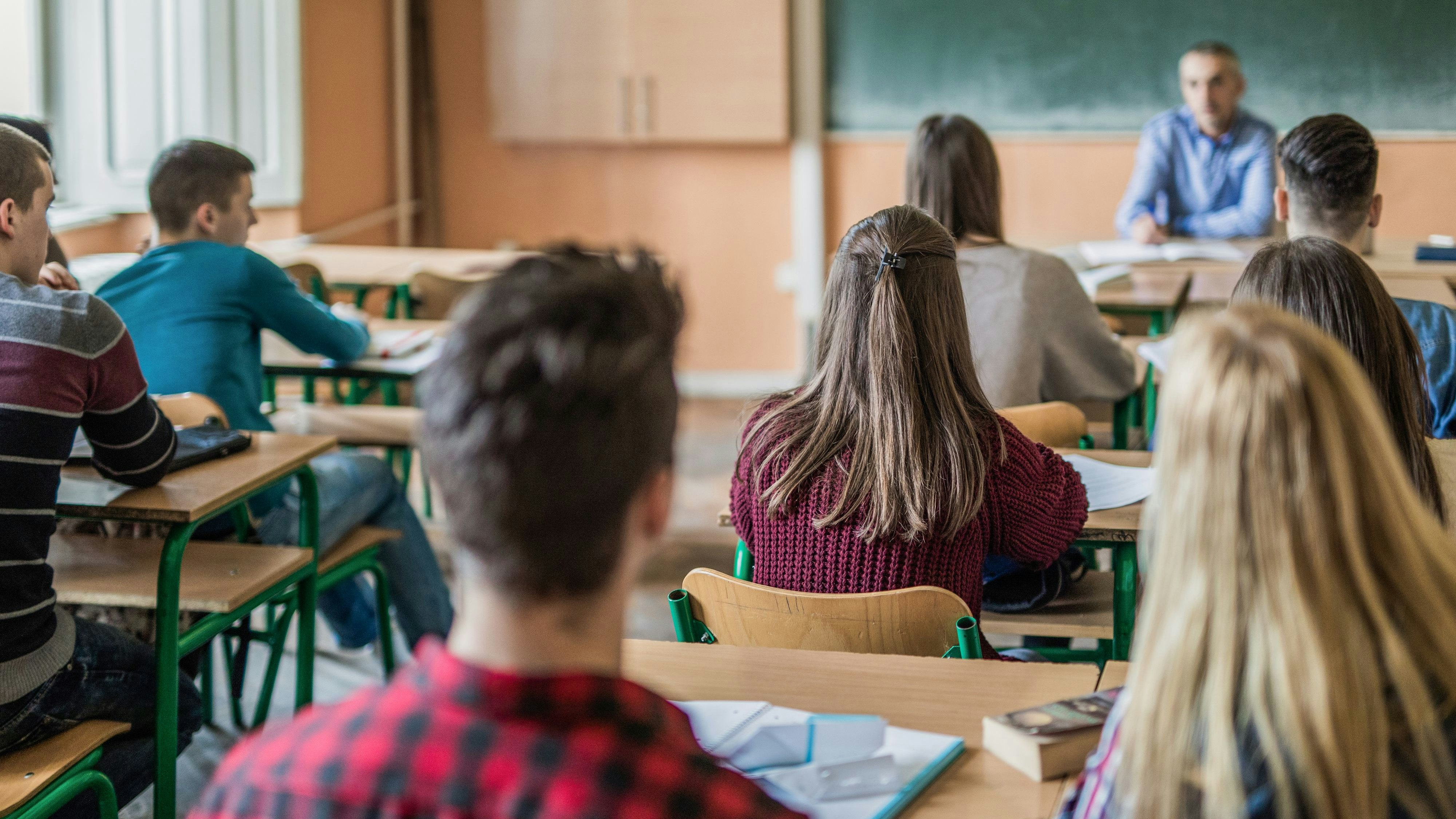 Back view of large group of students sitting in the classroom and listening to their teacher. Focus is on girl in purple sweater.