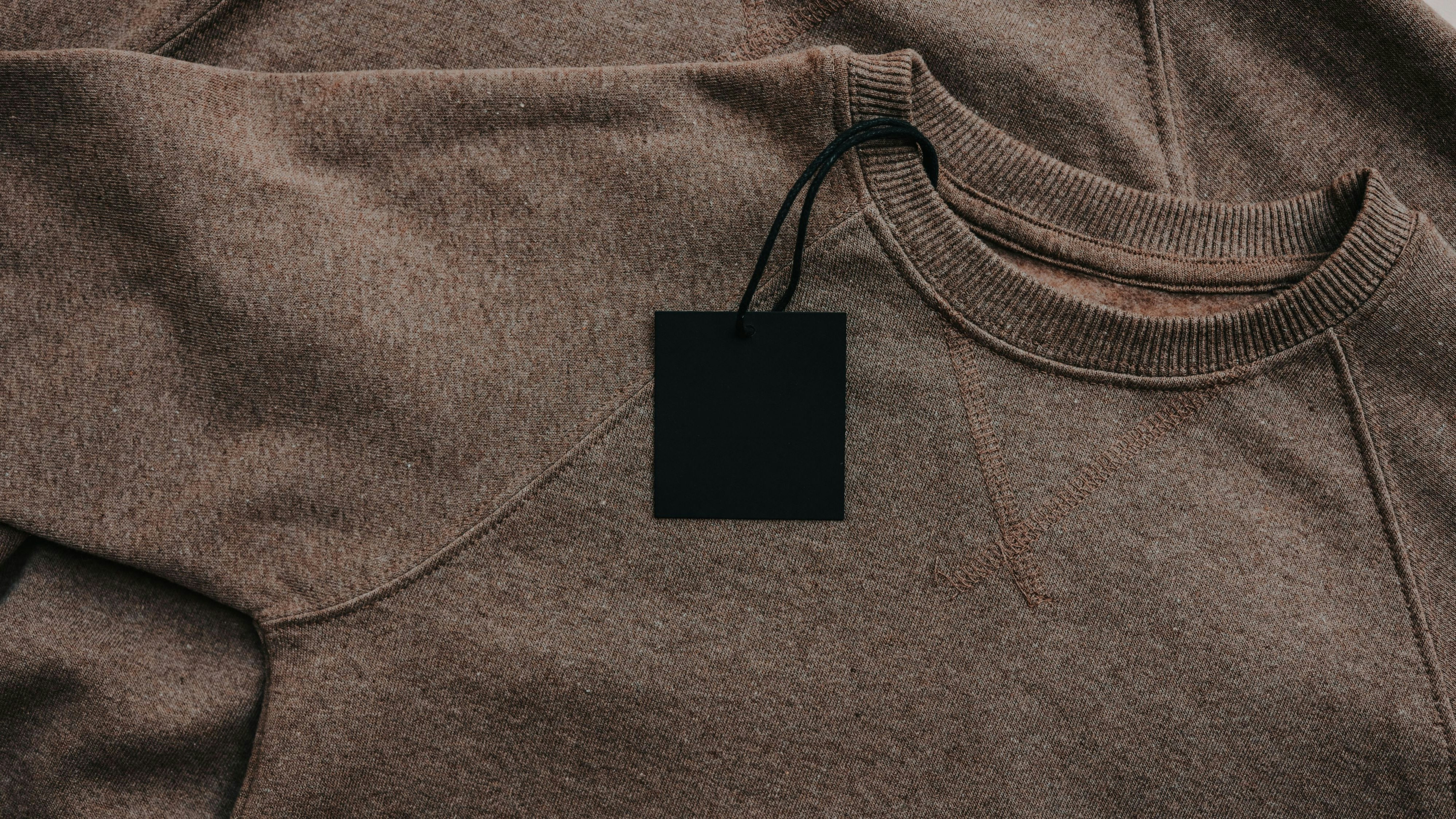 A close-up view of a soft brown crewneck sweatshirt featuring a plain black hang tag attached, highlighting the garment's material and construction