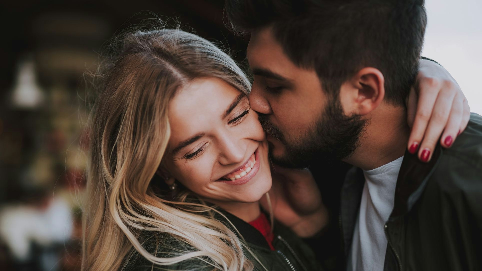 Precious moments of love. Close up portrait of handsome bearded guy kissing his girlfriend in cheek while she hugging him. Lady closing eyes with pleasure and smiling