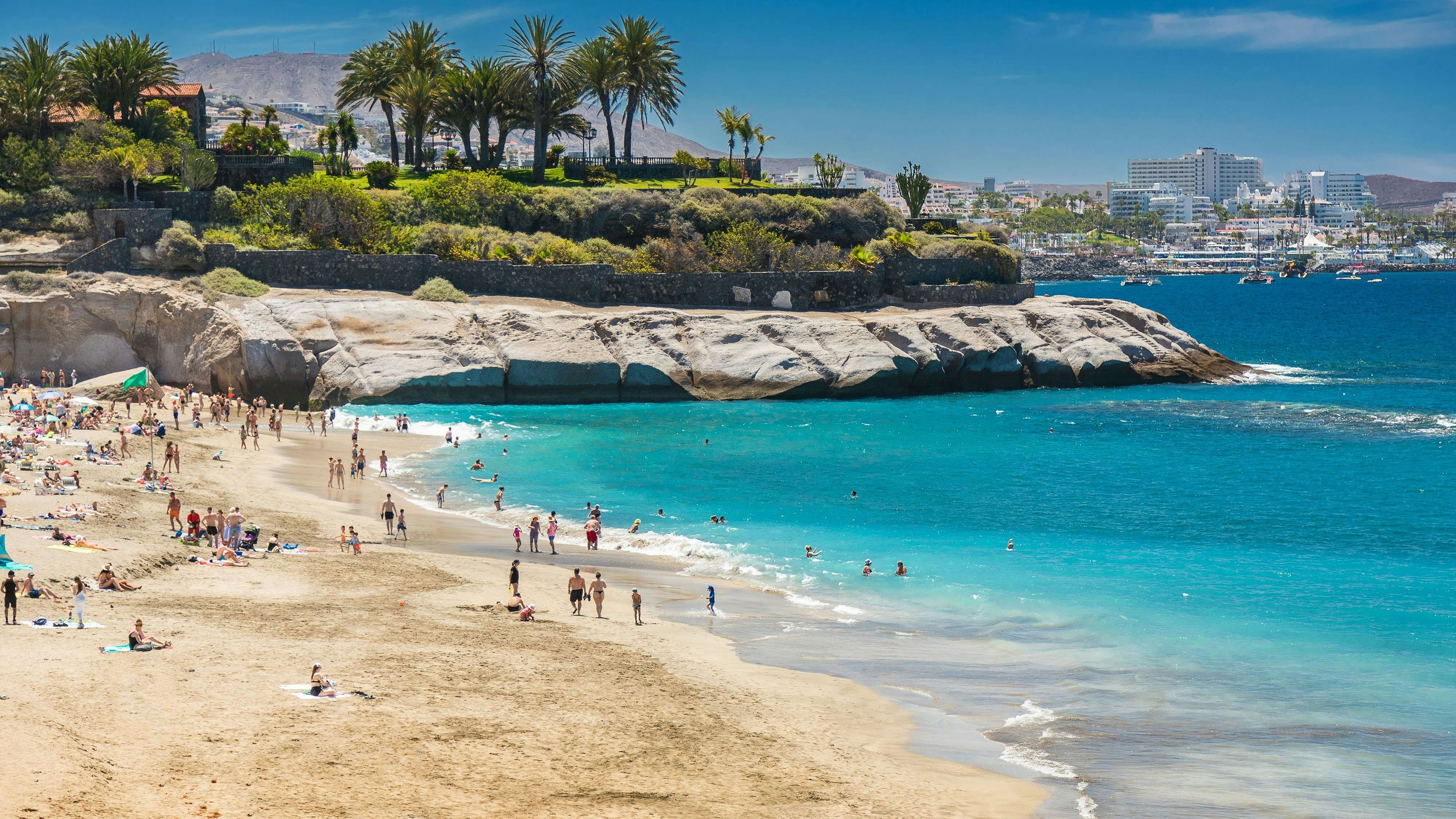 Playa del Duque offers a serene beach day with golden sand, clear blue water, and a bustling crowd enjoying the sun. Palm trees, rocky cliffs, and distant buildings add to the scenic charm of this Tenerife beach.