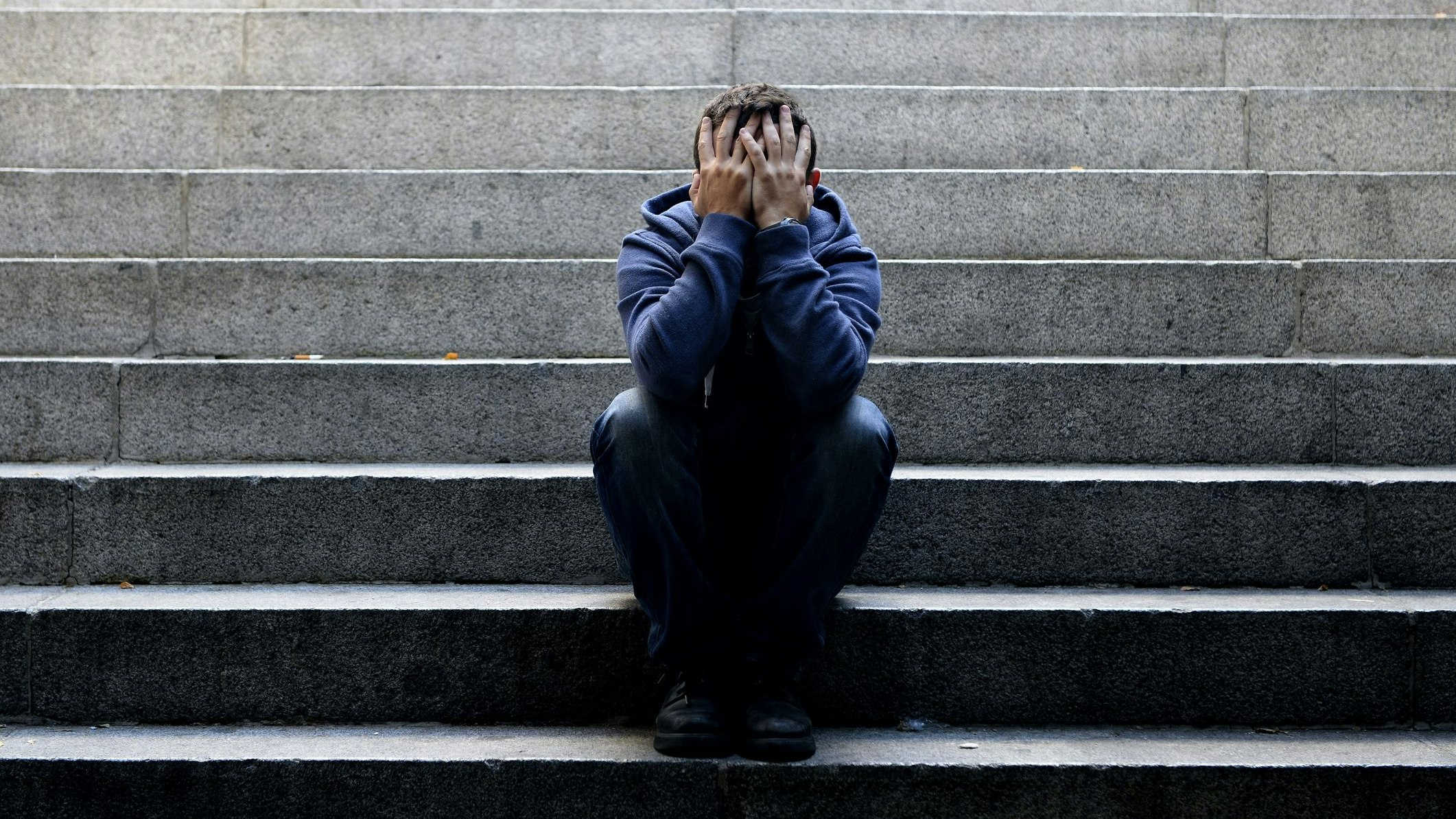 Young desperate man in casual clothes abandoned lost in depression sitting on ground street concrete stairs alone suffering emotional pain, sadness, looking sick in grunge lighting