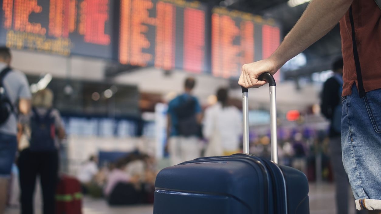 Traveling by airplane. Man waiting in airport terminal. Selective focus on hand holding suitcase against arrival and departure board. Passenger is ready for travel.