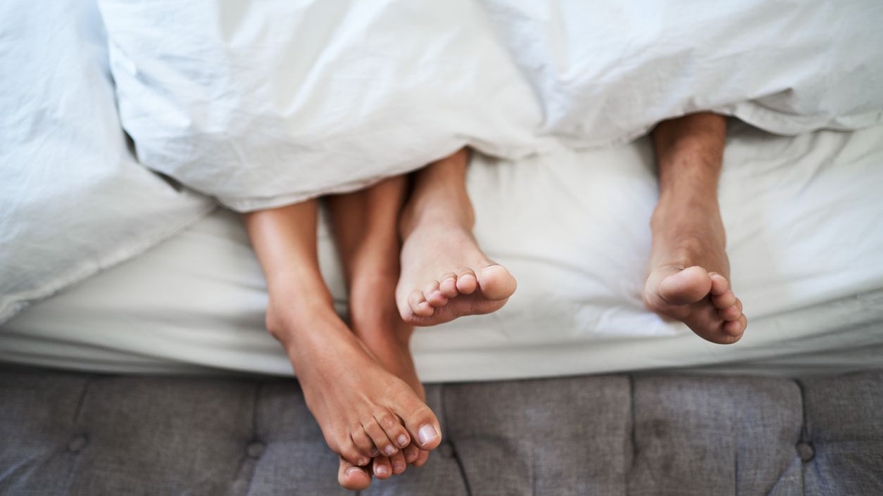 Two individuals' bare feet are shown sticking out from underneath a white duvet. This intimate scene depicts warmth, comfort, and a caring relationship, set within a cozy and private home environment.