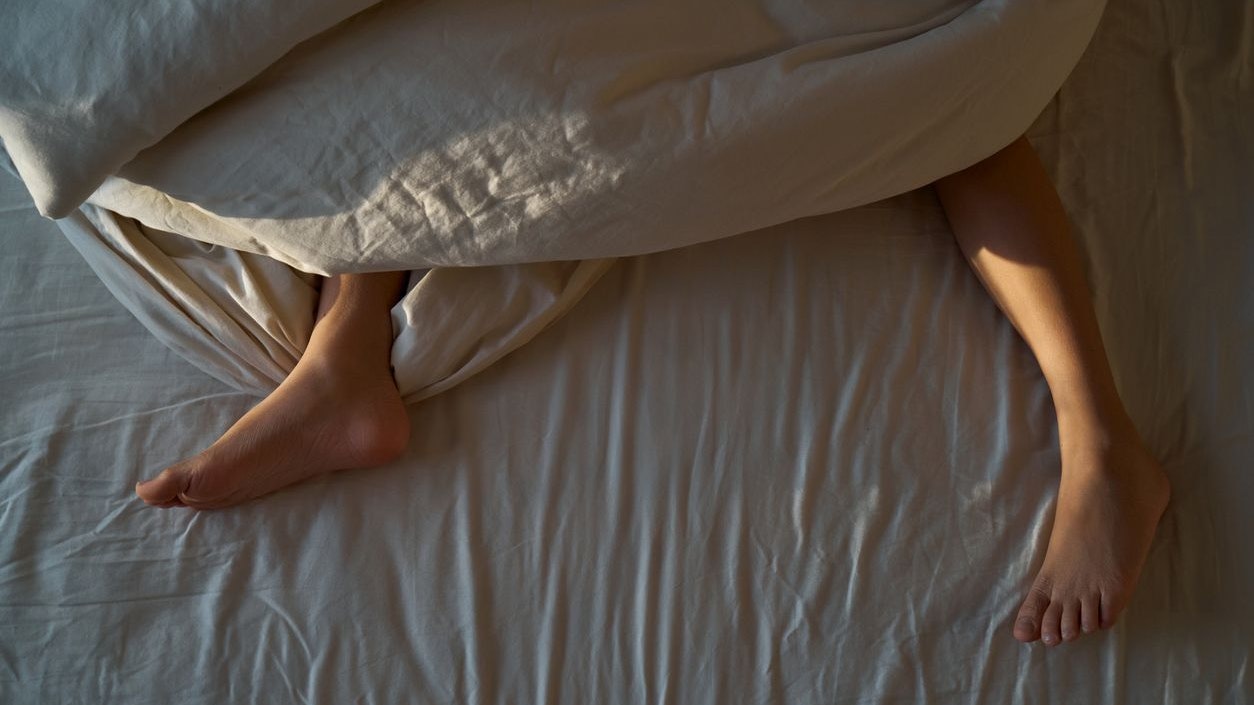 Person's feet and legs resting comfortably under a duvet in a bed, with morning sunlight creating a warm atmosphere