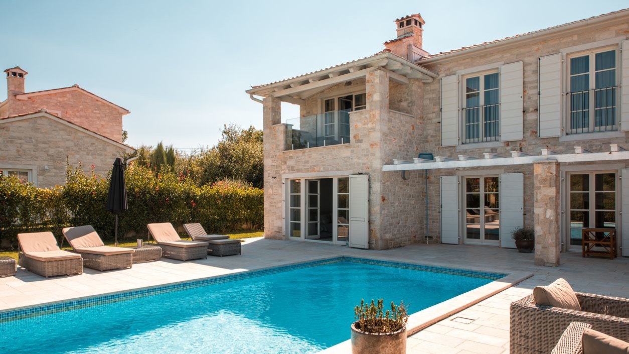 Wide angle shot of a stone villa in Mediterranean countryside. A large pool and sunbeds for multiple guests. Potted bush and wicker lounge chairs on the terrace in front of the house.