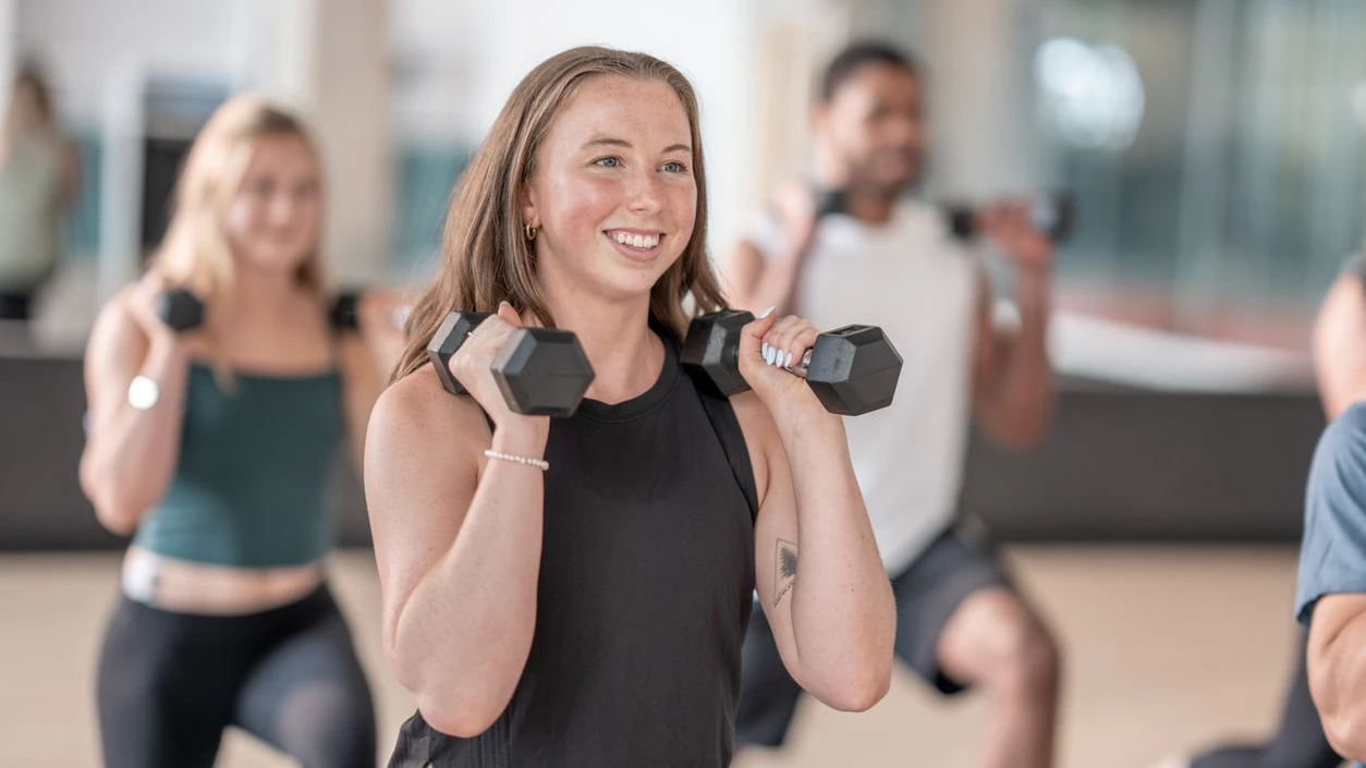 A group fitness class featuring individuals engaging in strength training exercises with dumbbells. Smiling participants showcase enthusiasm and teamwork in a modern gym environment promoting health and an active lifestyle.